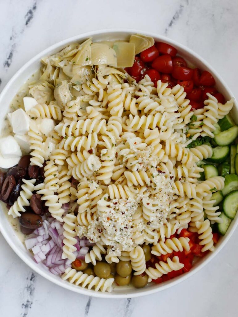 A bowl with ingredients for a pasta salad with dressing on top before mixing.