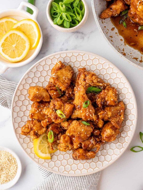 A white plate with Asian Lemon Chicken and a fresh lemon wedge. A bowl of fresh lemons is off to the side.