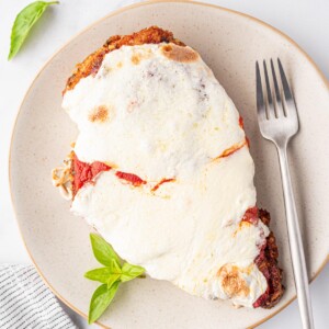 Oven Baked Chicken Parmesan is on an off-white plate with a silver fork and a few leaves of fresh basil.