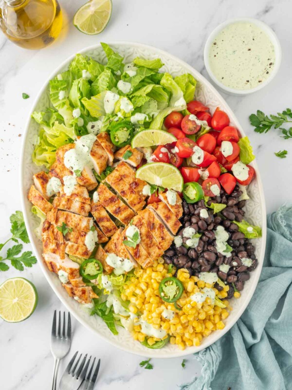 Ingredients for grilled chicken salad recipe piled on a plate with forks and dressing to the side.