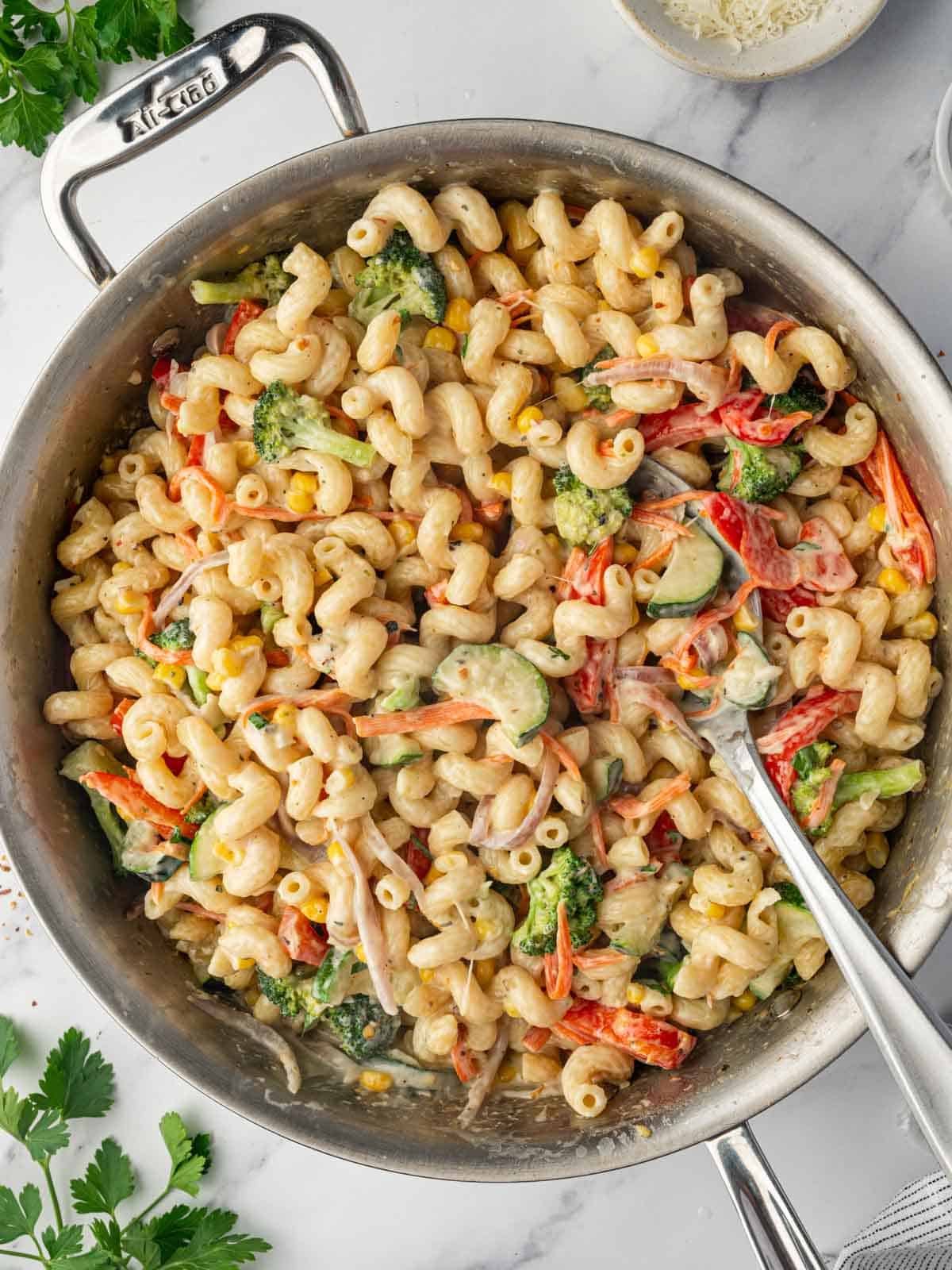 A spoon serves cheesy pasta with vegetable from a skillet.