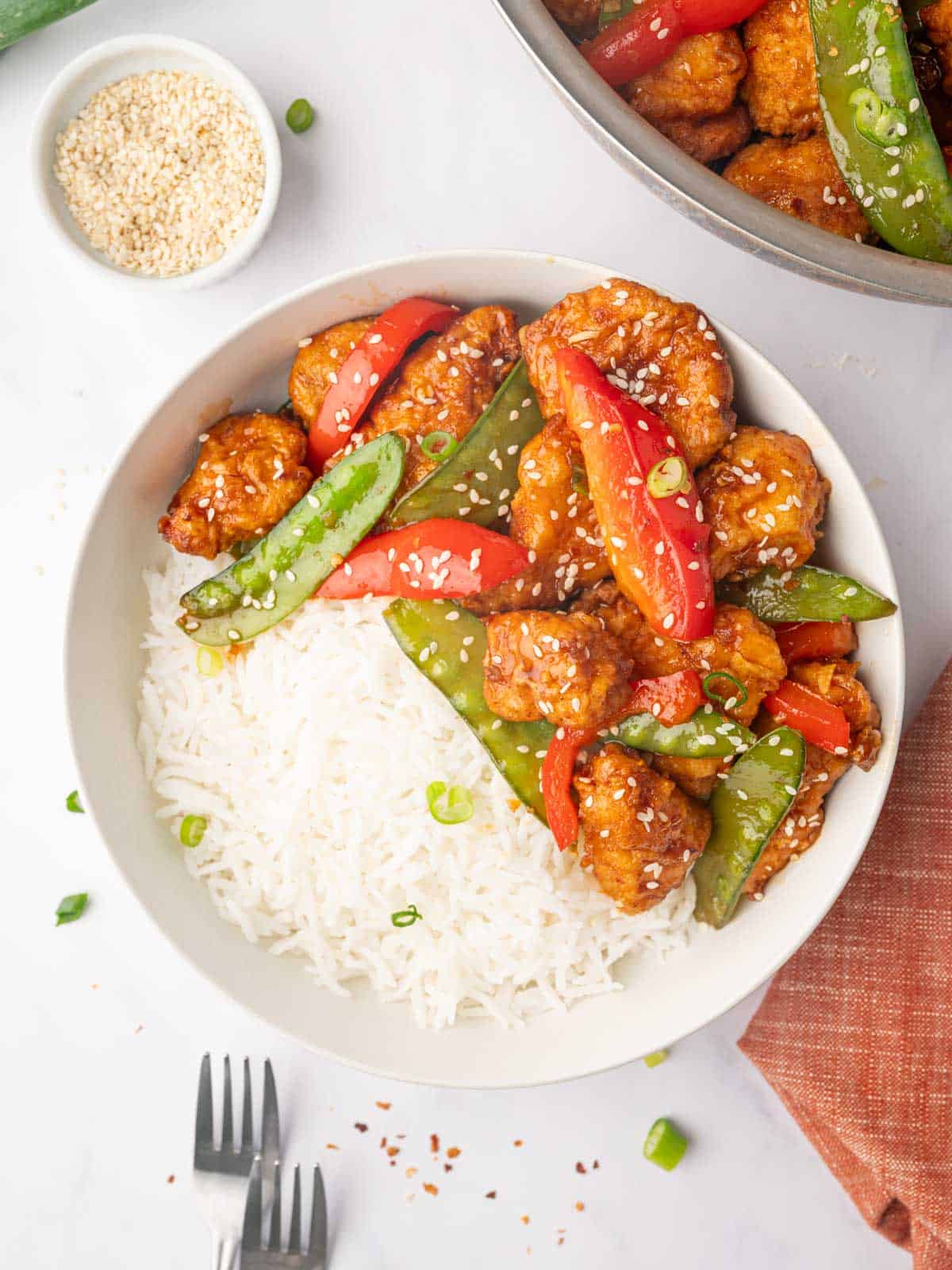 A plate with rice, veggies and crispy honey chili chicken garnished with sesame seeds.