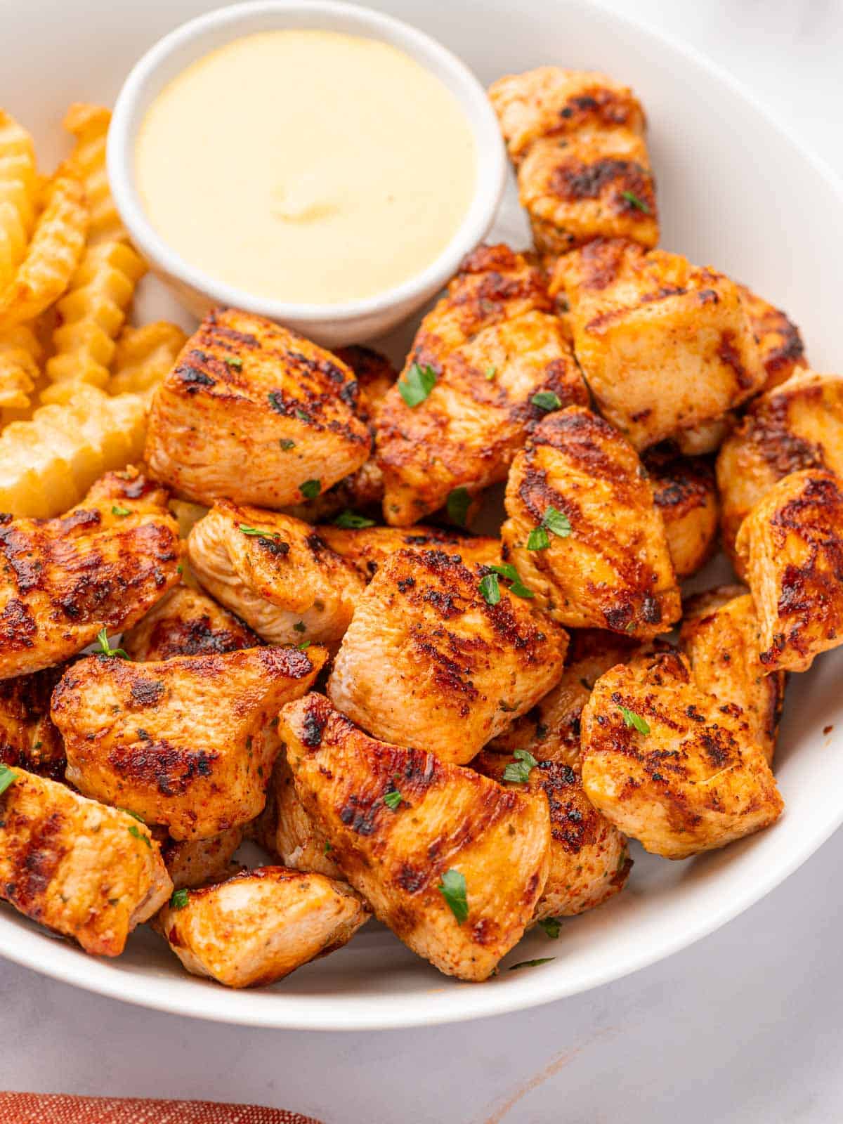 A pile of chicken nuggets with grill marks on a white plate with fries and a dipping sauce.