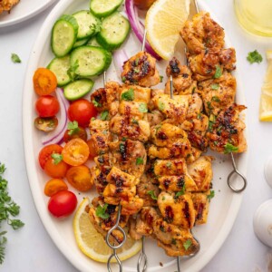 Fresh veggies and lemon wedges surround grilled chicken skewers on a platter.